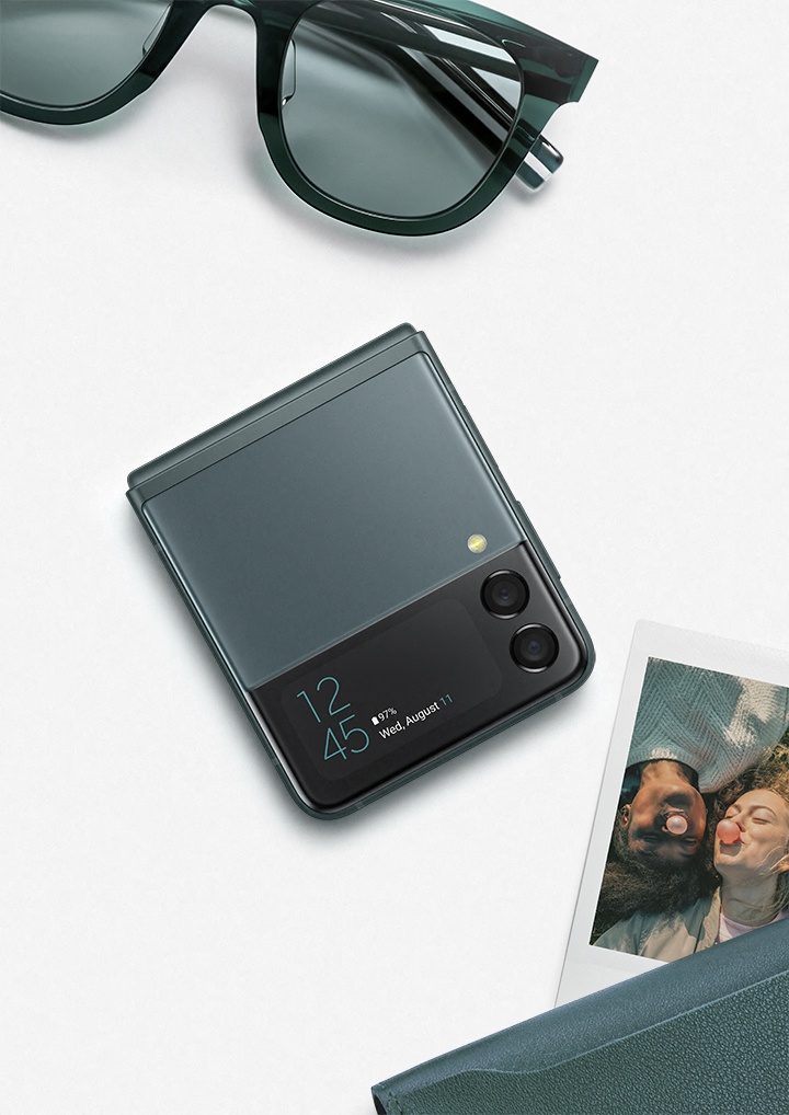 Óculos de sol pretos, Samsung Galaxy Z Flip3 5G dobrado e uma fotografia polaroid com uma selfie de duas pessoas em cima de uma superfície branca